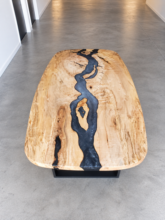 Custom squoval light-wood dining table with a dark resin river and black metal base in a clean showroom.
